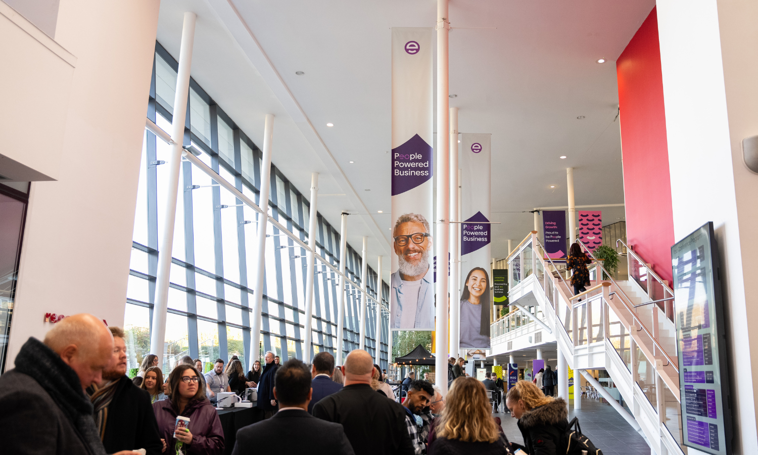 A gathering of people inside a glass fronted building. Banners promoting the eoa hang down from the ceiling.