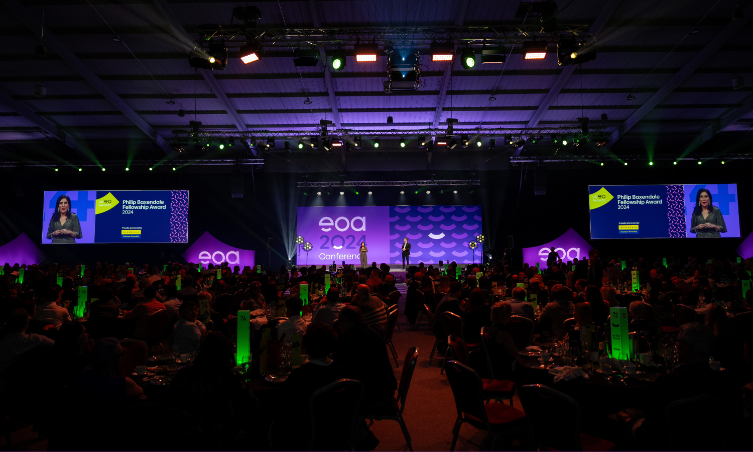 An business awards ceremony with two people on stage and tables gathered in cabaret style in front.